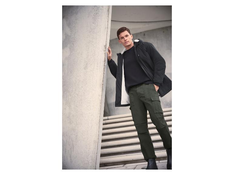 Muž v čiernej bunde s bielou podšívkou, čiernom tričku a zelených cargo nohaviciach.