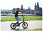 Žena jazdí na elektrobicykli Sachsengrad po nábreží s panorámou mesta v pozadí.