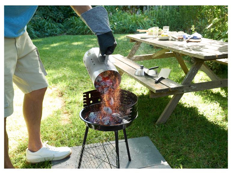 Muž sype žeravé uhlie z podpaľovacieho komína do grilu, vedľa piknikový stôl s riadom.