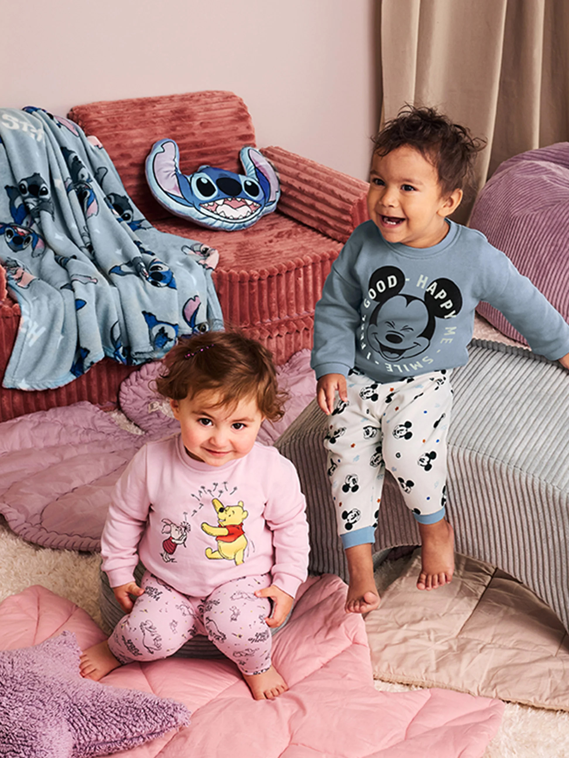 Dve bábätká v pyžamách Disney, obklopené prikrývkami a vankúšmi Stitch.