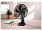 Čierny stolný ventilátor na drevenom stole s poznámkovými blokmi a perami, vo svetlej miestnosti.
