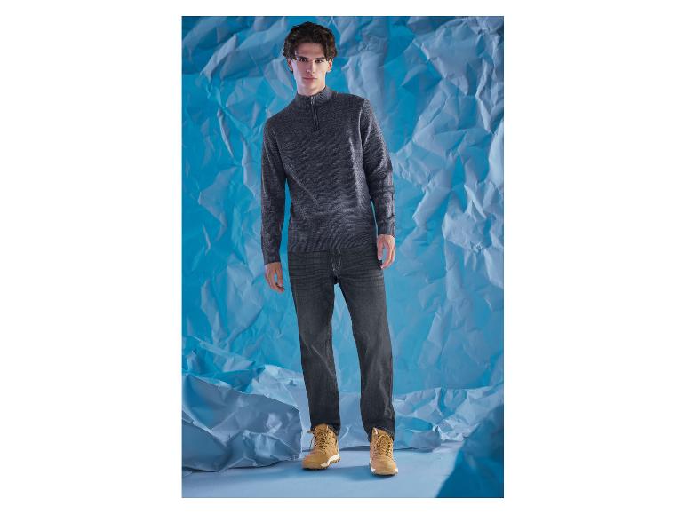 Muž v sivom svetri a tmavých džínsoch stojí pred modrým pozadím.