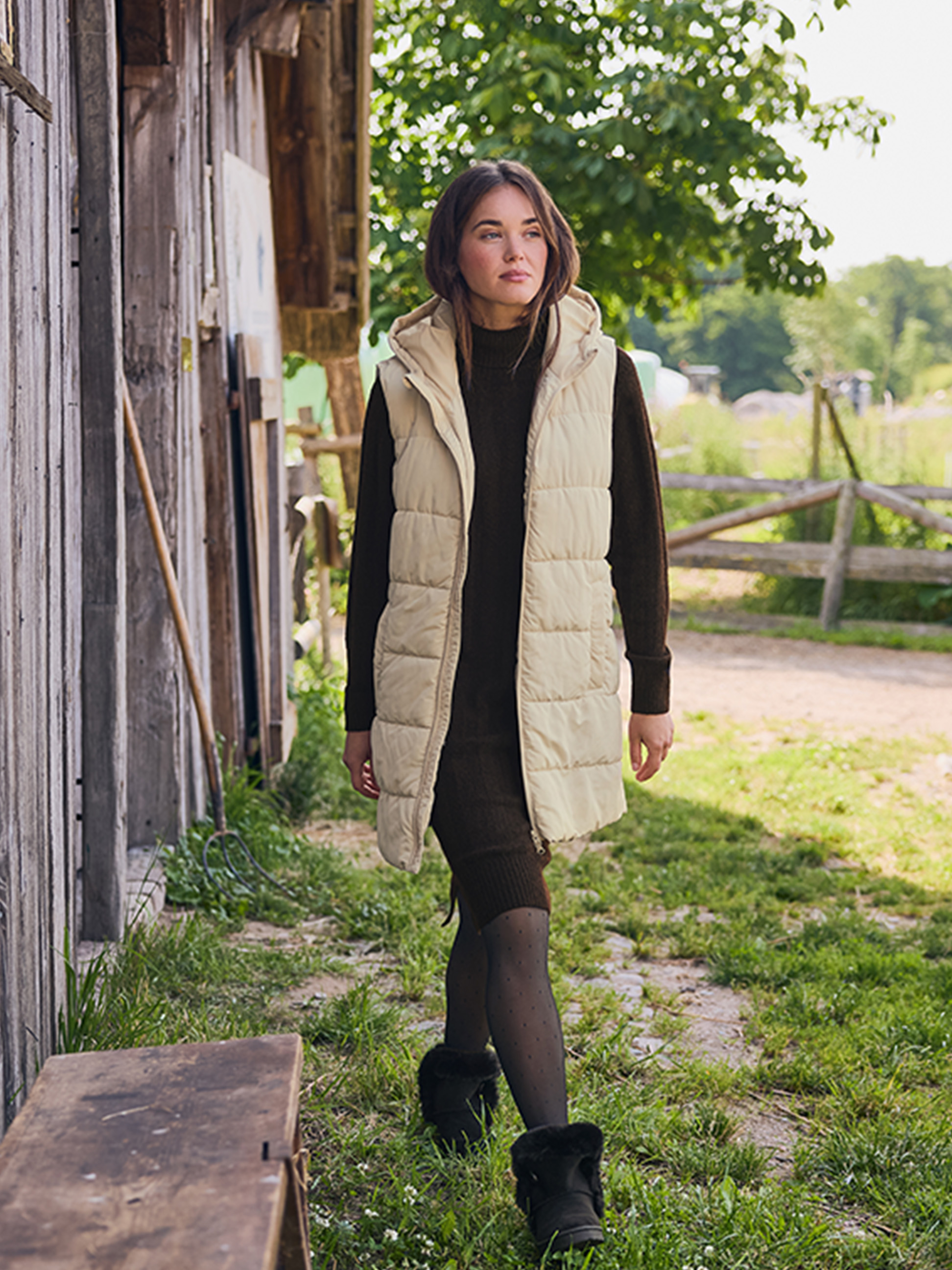 Žena v béžovej veste a hnedých šatách, kráčajúca po vidieckej ceste.