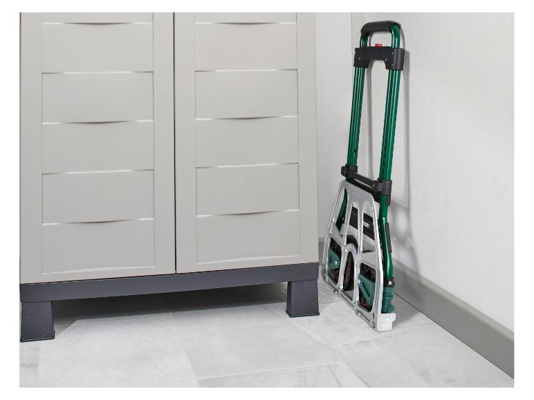 Zložený zelený a strieborný ručný vozík opretý o bielu stenu vedľa svetlosivej úložnej skrinky.