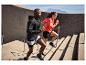 Dvaja športovci v aktívnom oblečení bežia po vonkajších schodoch s horami v pozadí.