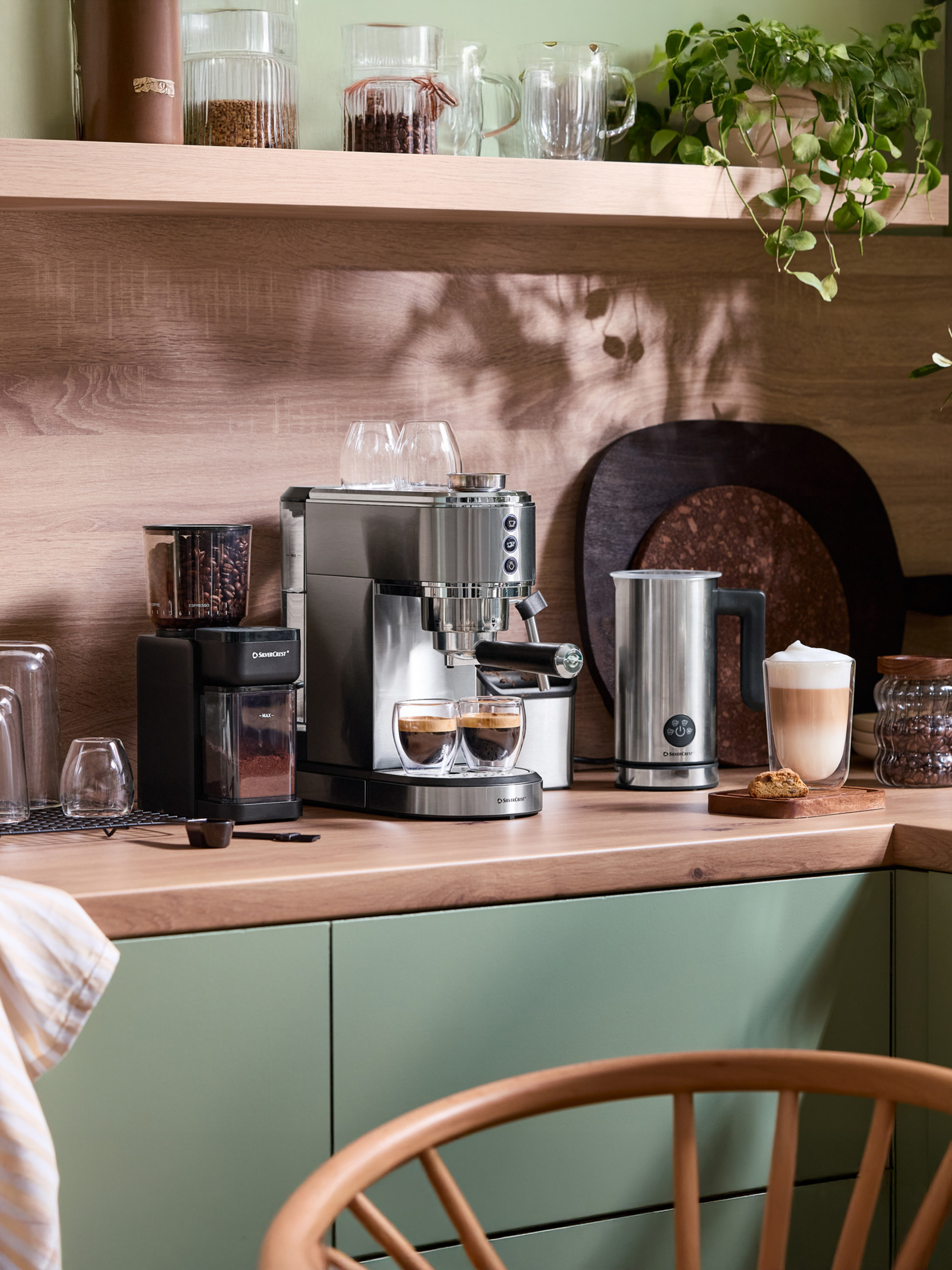 Kávovar SilverCrest a mlynček na kávu na drevenej kuchynskej linke so zelenými skrinkami.