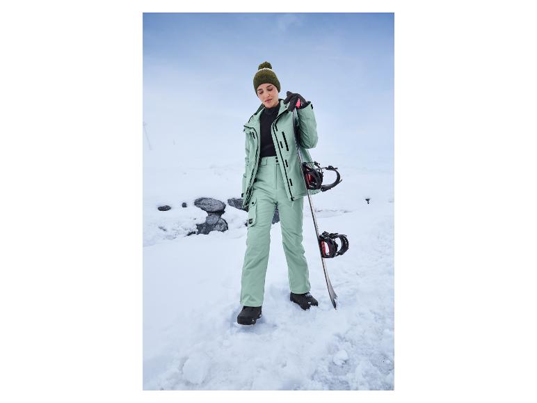 Žena v zelených lyžiarskych nohaviciach a bunde s snowboardom v snehu.