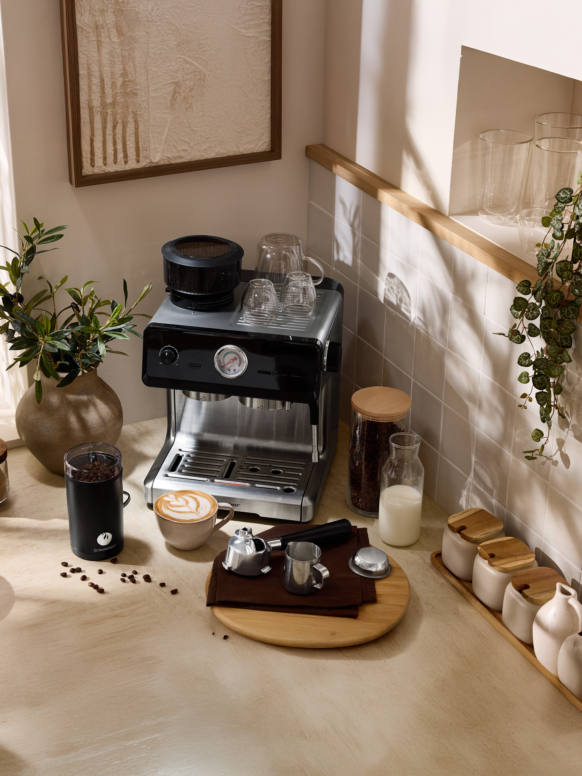 Kávovar s mlynčekom, šálkou latte kávy a príslušenstvom na kuchynskej linke.