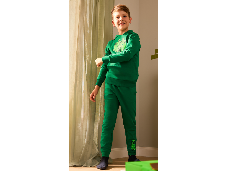 Usmiaty chlapec v zelenej mikine s kapucňou a teplákoch s potlačou príšery.