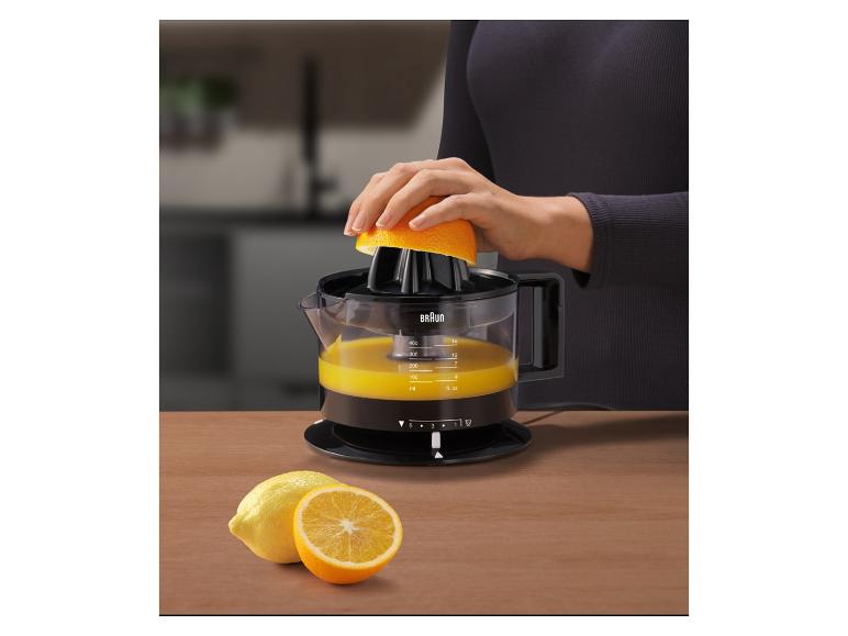 Žena odšťavuje pomaranč citrusovačom, s citrónmi a pomarančmi na drevenom stole.