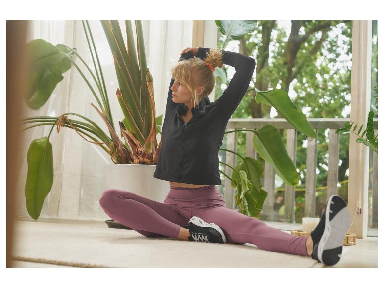 Žena v čiernej športovej bunde a fialových legínach sa naťahuje v interiéri.