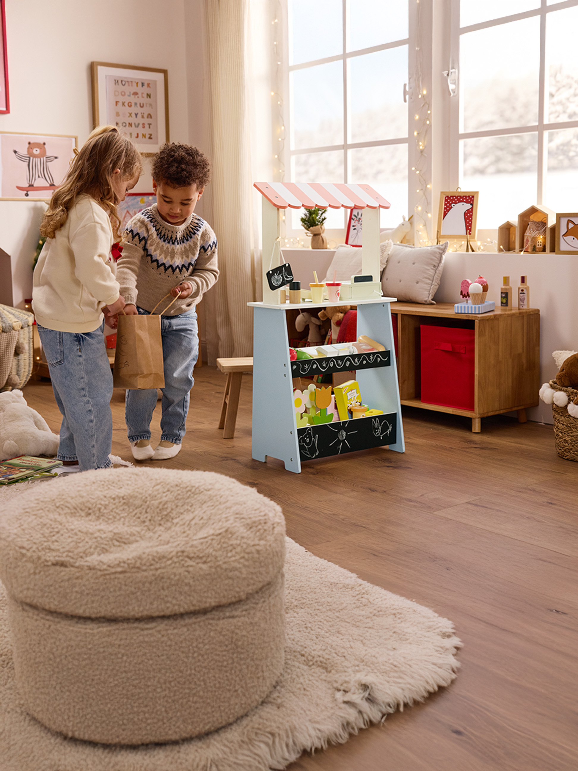 Dve deti sa hrajú s dreveným trhovým stánkom, dievča drží papierovú tašku.