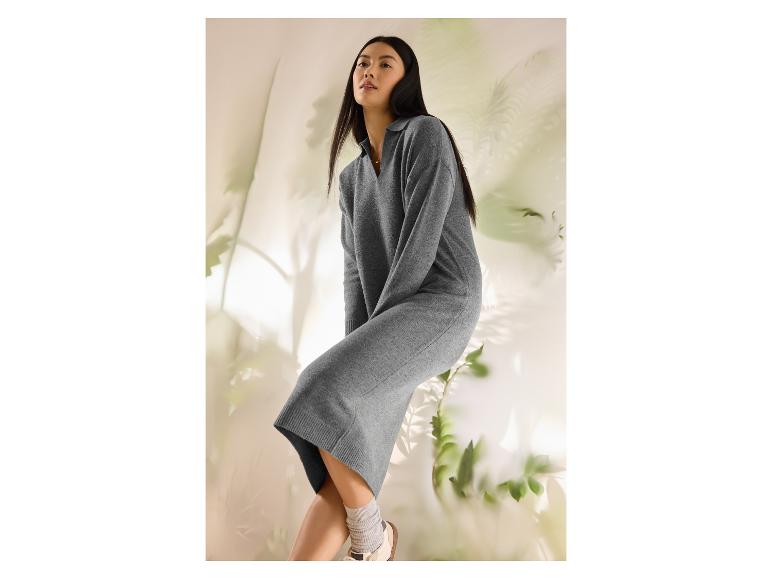 Žena v sivých pletených šatách a bielych teniskách, sediaca na svetlom pozadí s rastlinami.