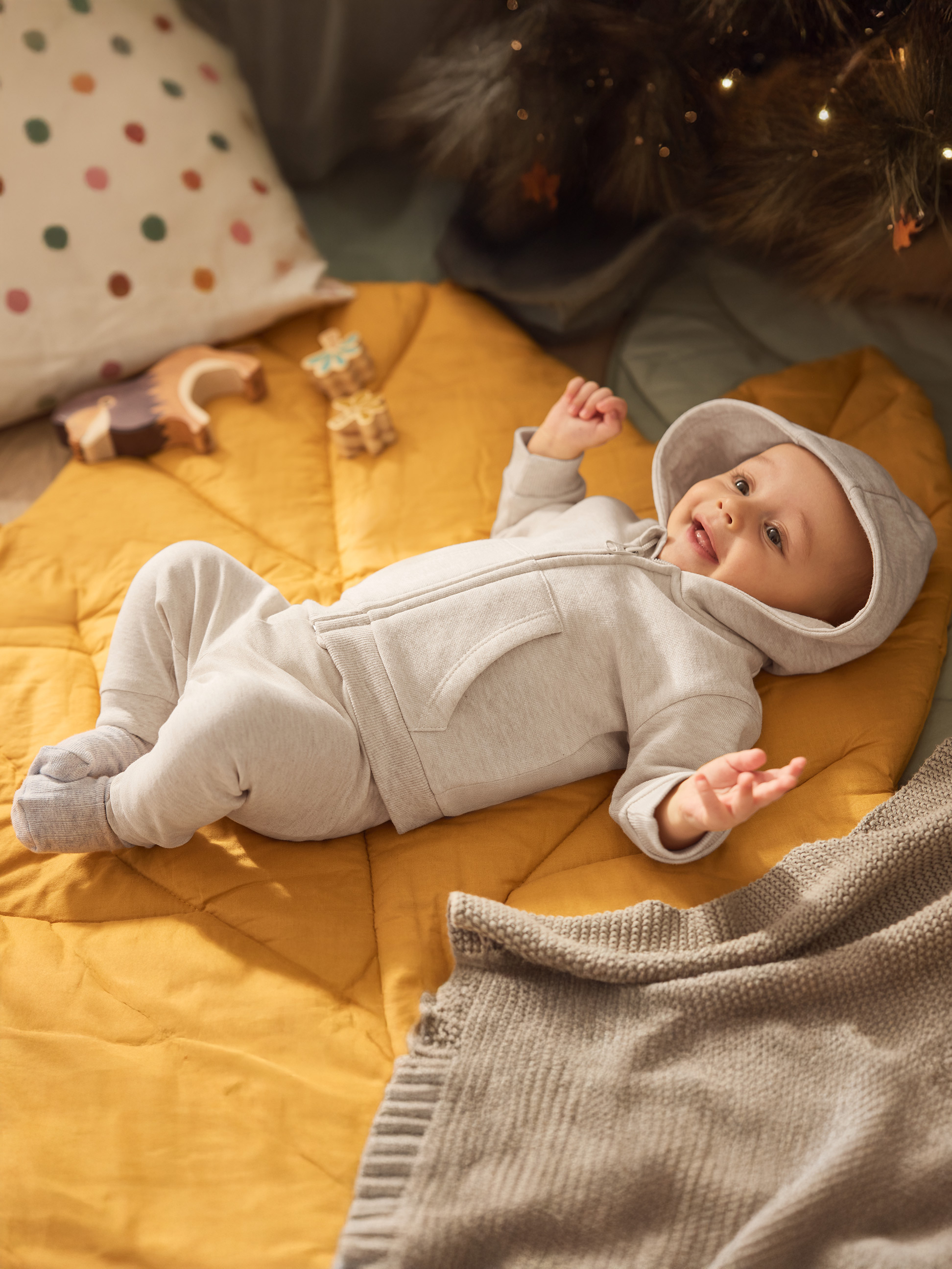 Šťastné bábätko v sivom overale na žltom matraci s hračkami a vianočnými svetielkami.