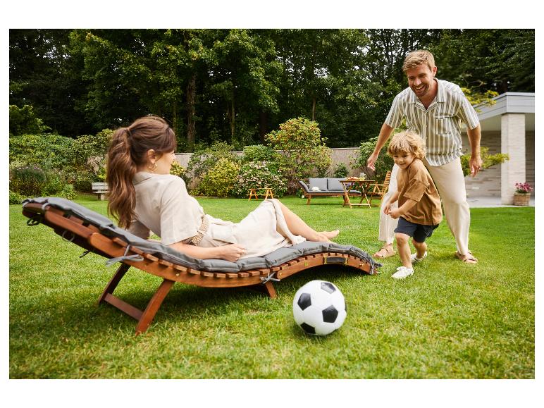 Žena relaxuje na drevenom ležadle so sivým vankúšom, dieťa a muž hrajú futbal.