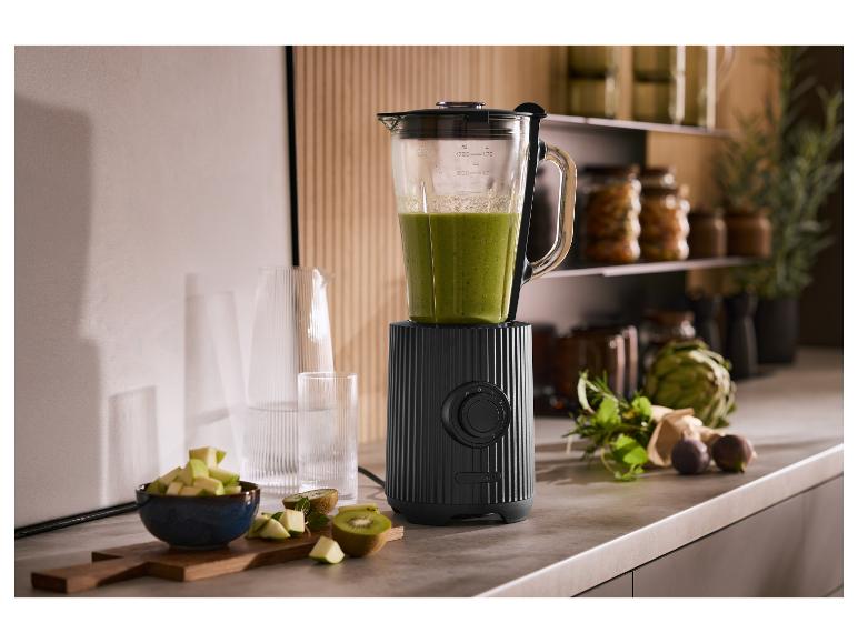 Čierny mixér so zeleným smoothie, ovocím a bylinkami v kuchyni.