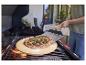 Pizza na pizza kameni na grile, s osobou používajúcou lopatu na pizzu.