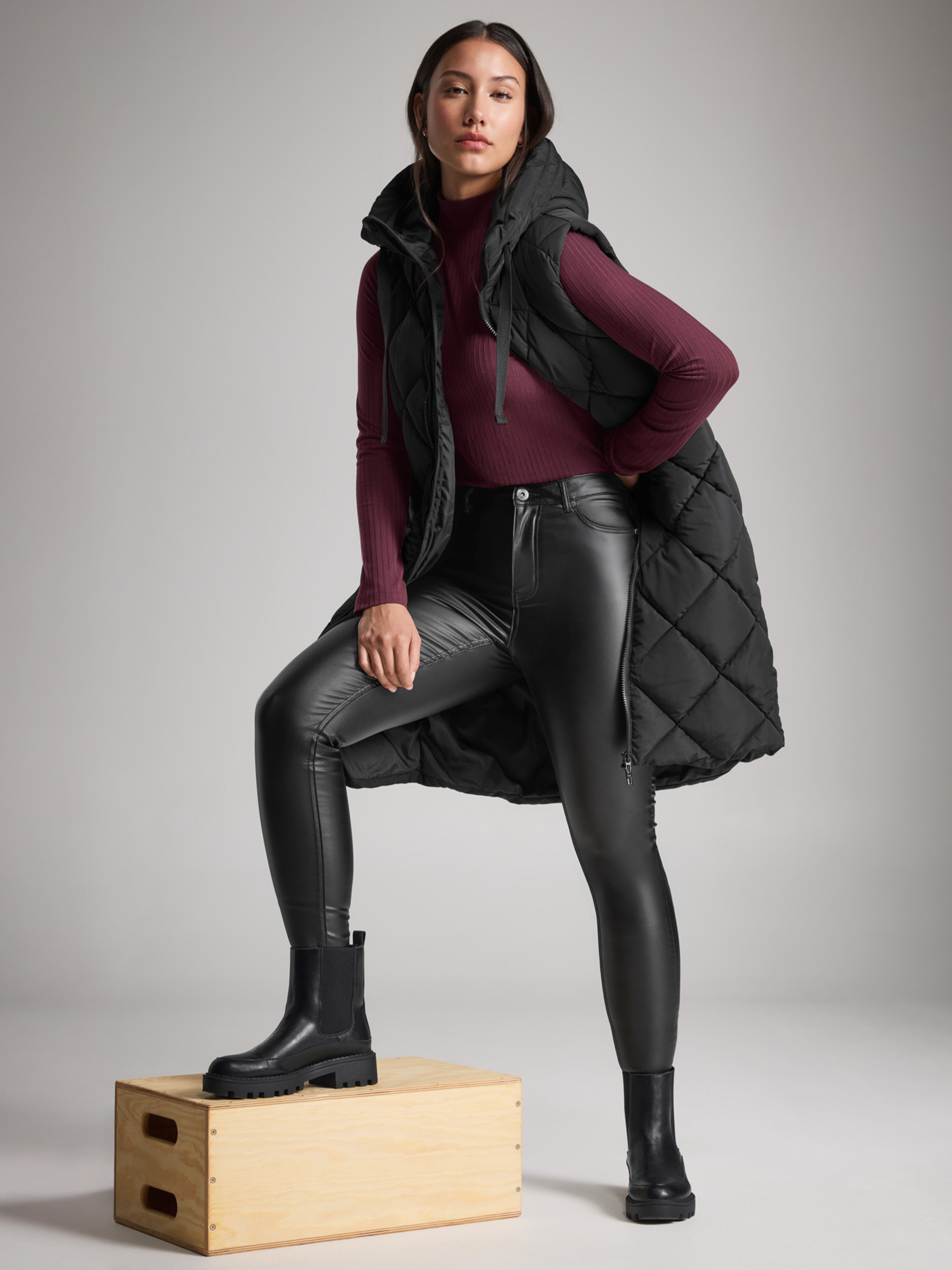 Žena v čiernej prešívanej veste, kožených džínsoch a čižmách, s jednou nohou na krabici.