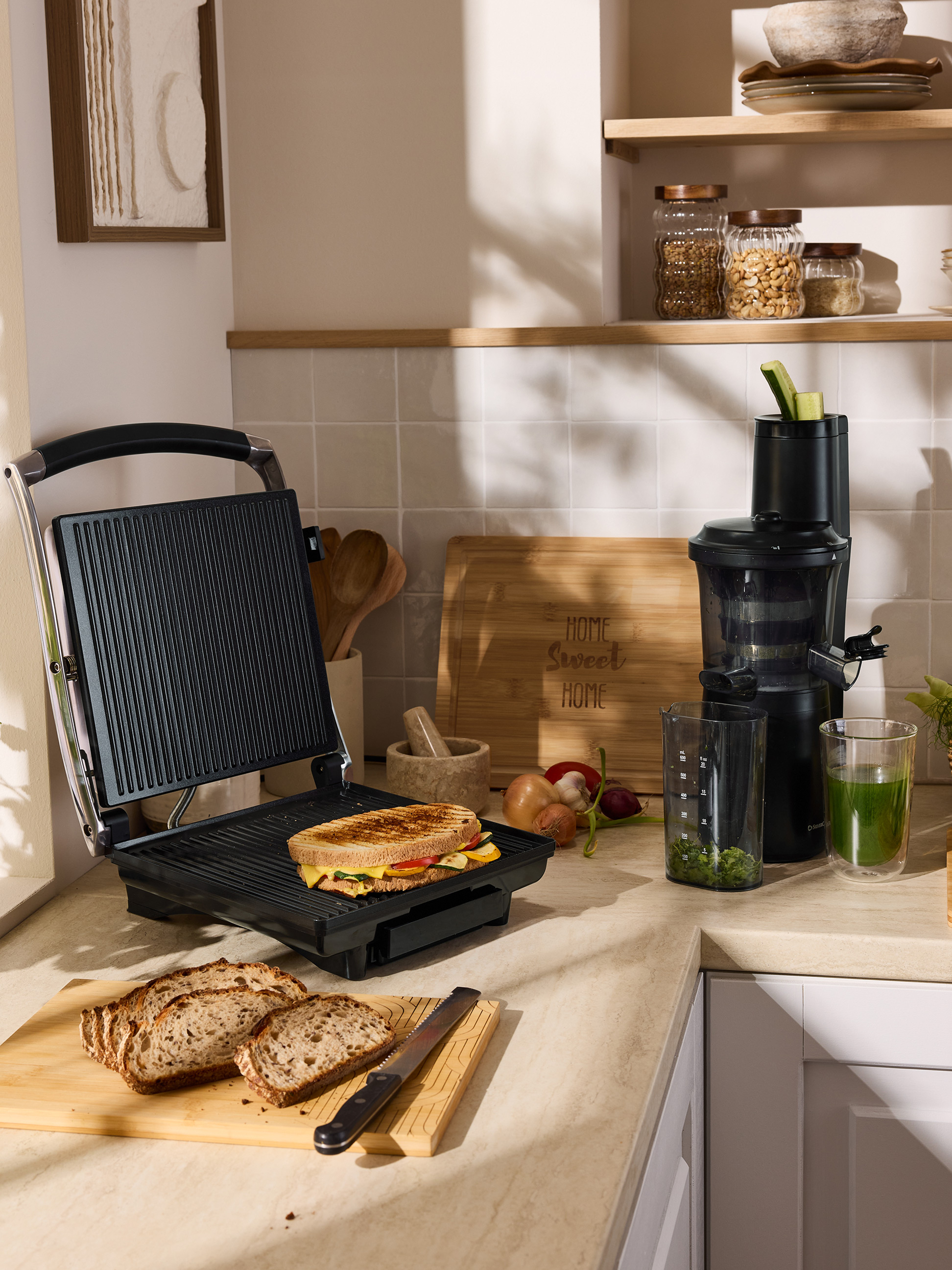 Grilovaný sendvič na sendvičovači, nakrájaný chlieb a odšťavovač na kuchynskej linke.