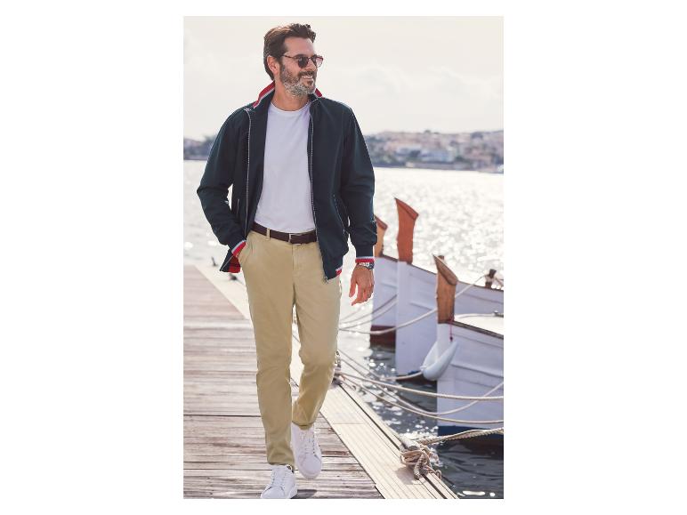 Muž v modrej bunde, béžových nohaviciach a bielych teniskách na móle.