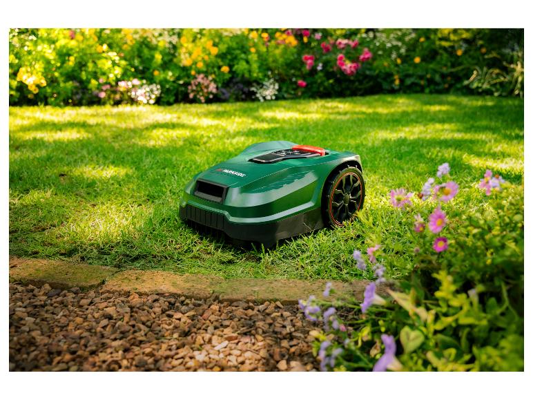 Zelená robotická kosačka na trávniku s kvetmi a kríkmi v pozadí.