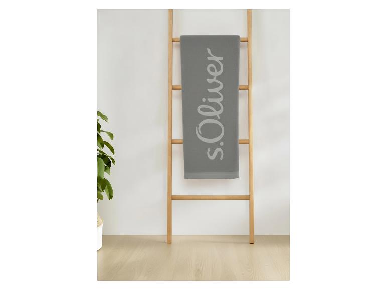 Sivý uterák S.Oliver s logom, zavesený na drevenom rebríku, vedľa rastliny.