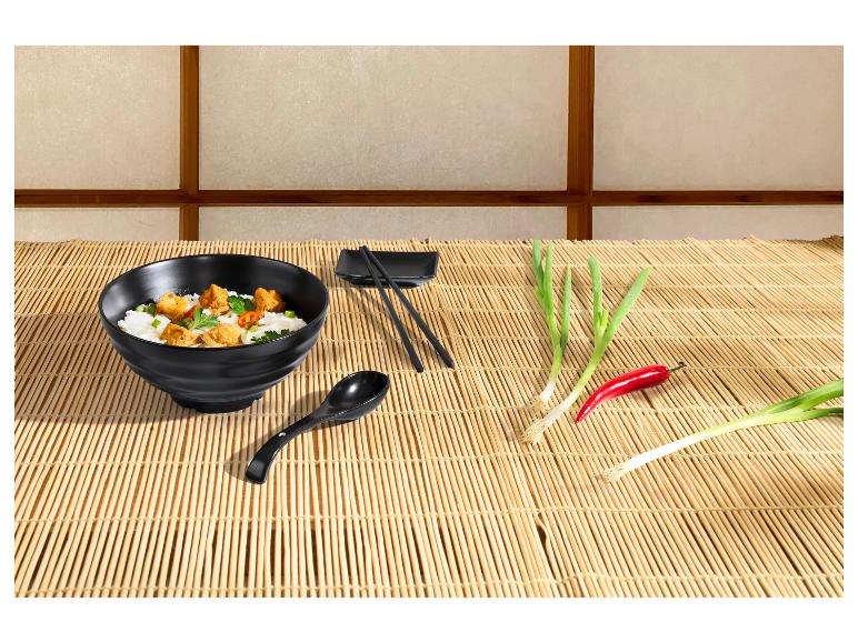 Čierna miska s ázijským jedlom, paličky, lyžica, jarná cibuľka a čili na bambusovej podložke.