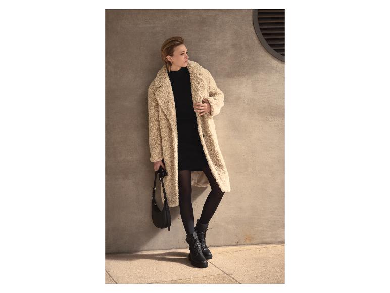 Žena v krémovom plyšovom kabáte, čiernych šatách, pančuchách a čižmách, držiaca čiernu tašku.