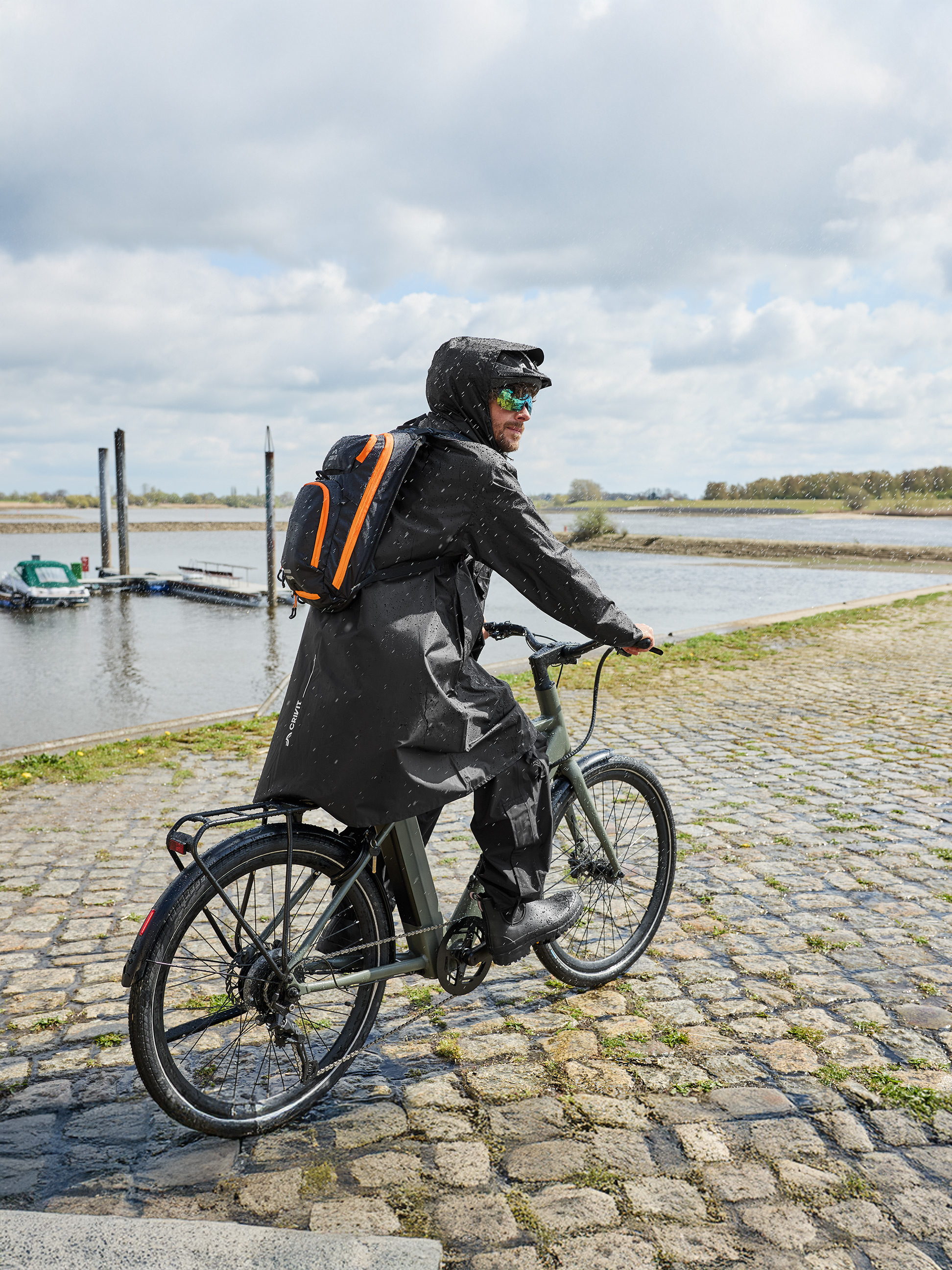 Muž v pršiplášti a s batohom jazdí na bicykli v daždi pri rieke.