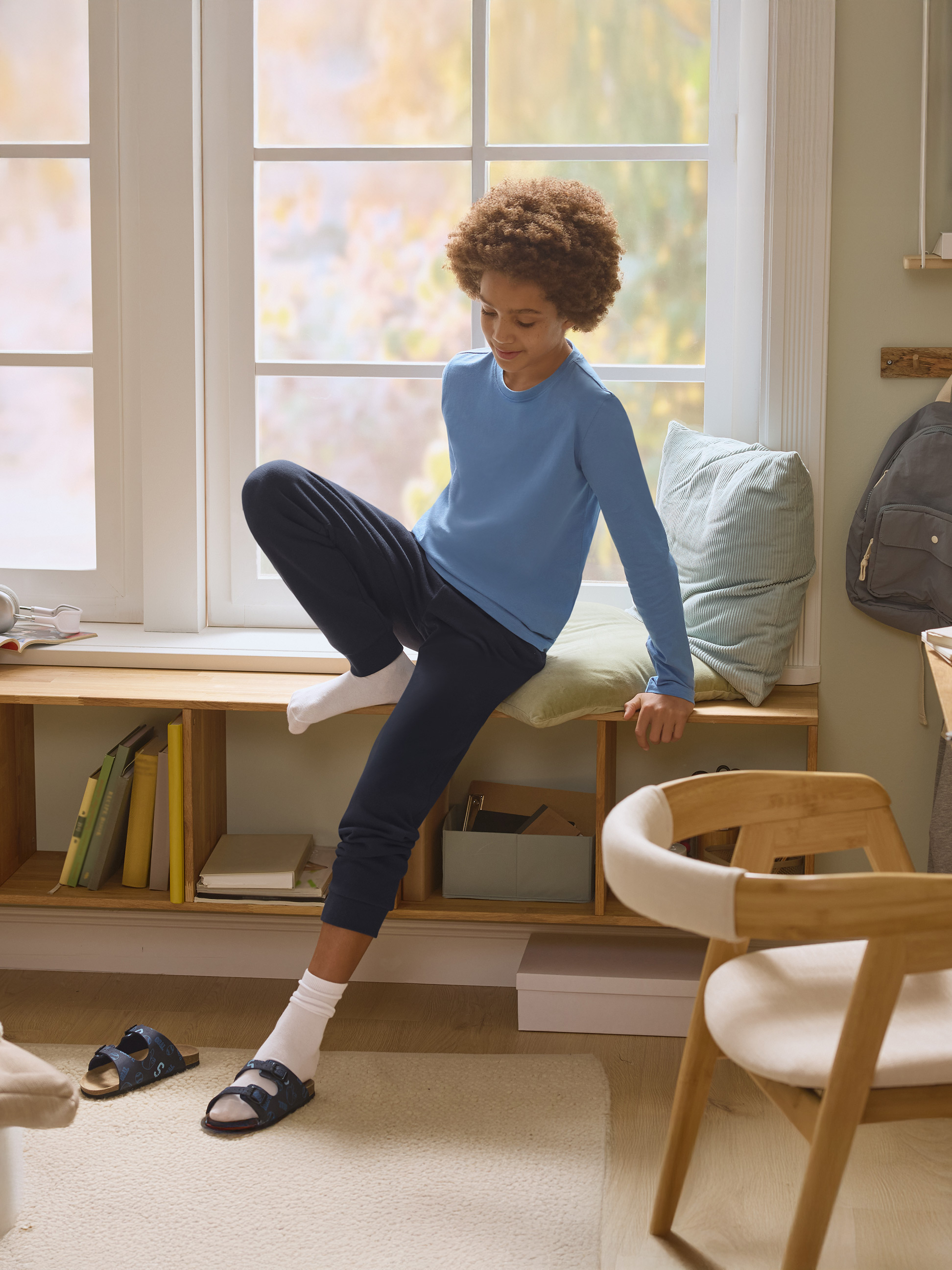 Chlapec v modrom pyžame sedí na parapete s knihami a vankúšmi.