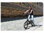 Žena na e-bicykli pred historickou nástennou maľbou, slnečný deň.