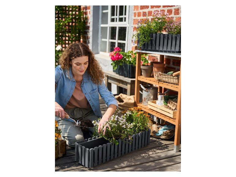 Žena sadí kvety do sivého kvetináča na drevenej terase, s záhradnou policou.