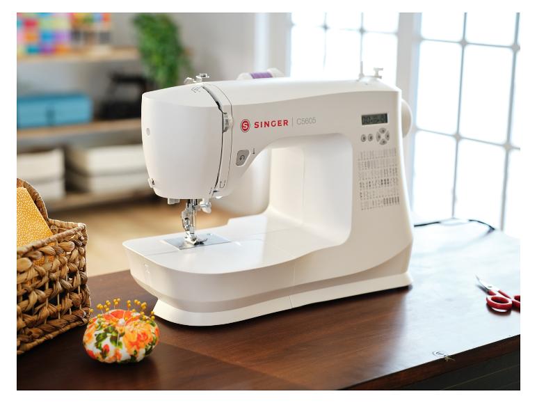 Biely šijací stroj Singer na drevenom stole, s košíkom potrieb a vankúšikom na ihly.