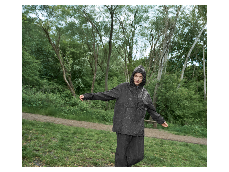 Žena v tmavom dažďovom kabáte a nohaviciach si užíva dážď v parku.