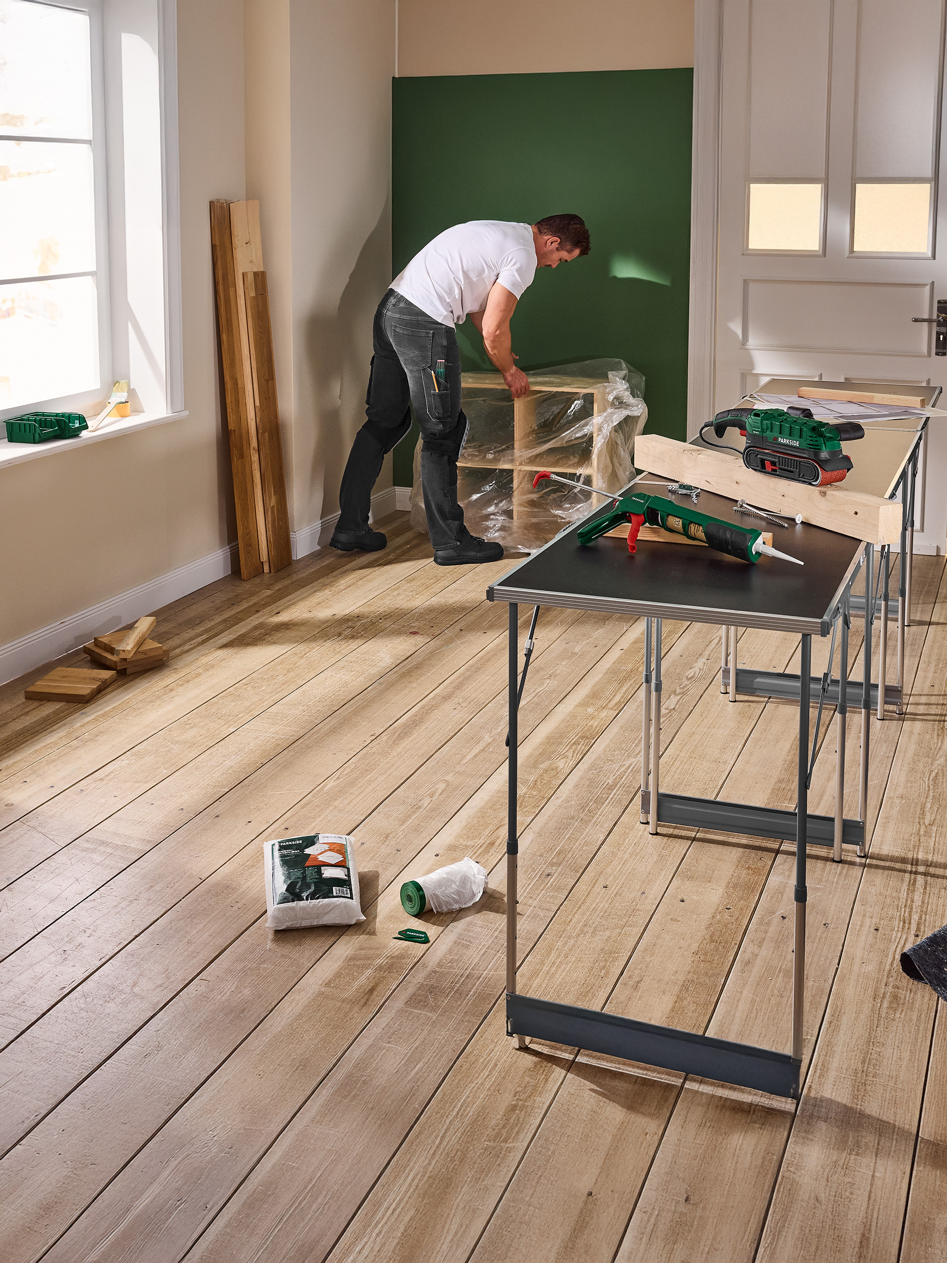 Muž pracuje na DIY projekte s náradím Parkside, vrátane brúsky a pištole na tmel, na skladacích stoloch.