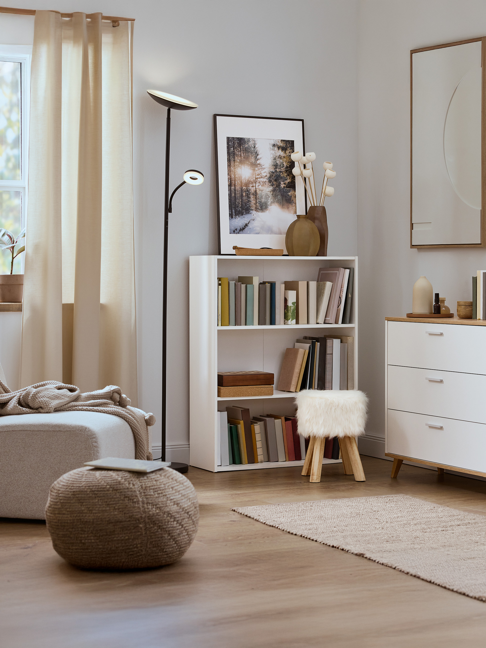 Svetlá obývacia izba s bielou knižnicou, pleteným pufom a modernou stojacou lampou.