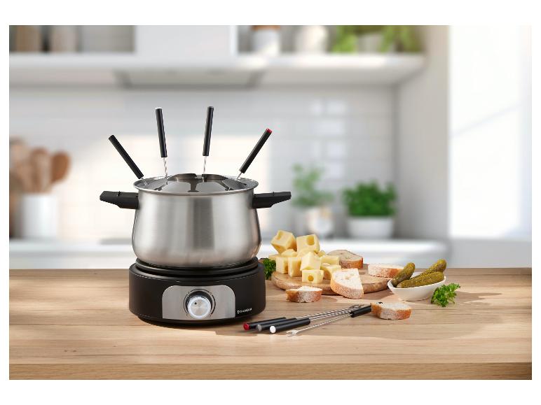 Fondue súprava so syrom, chlebom a uhorkami na drevenom stole v kuchyni.