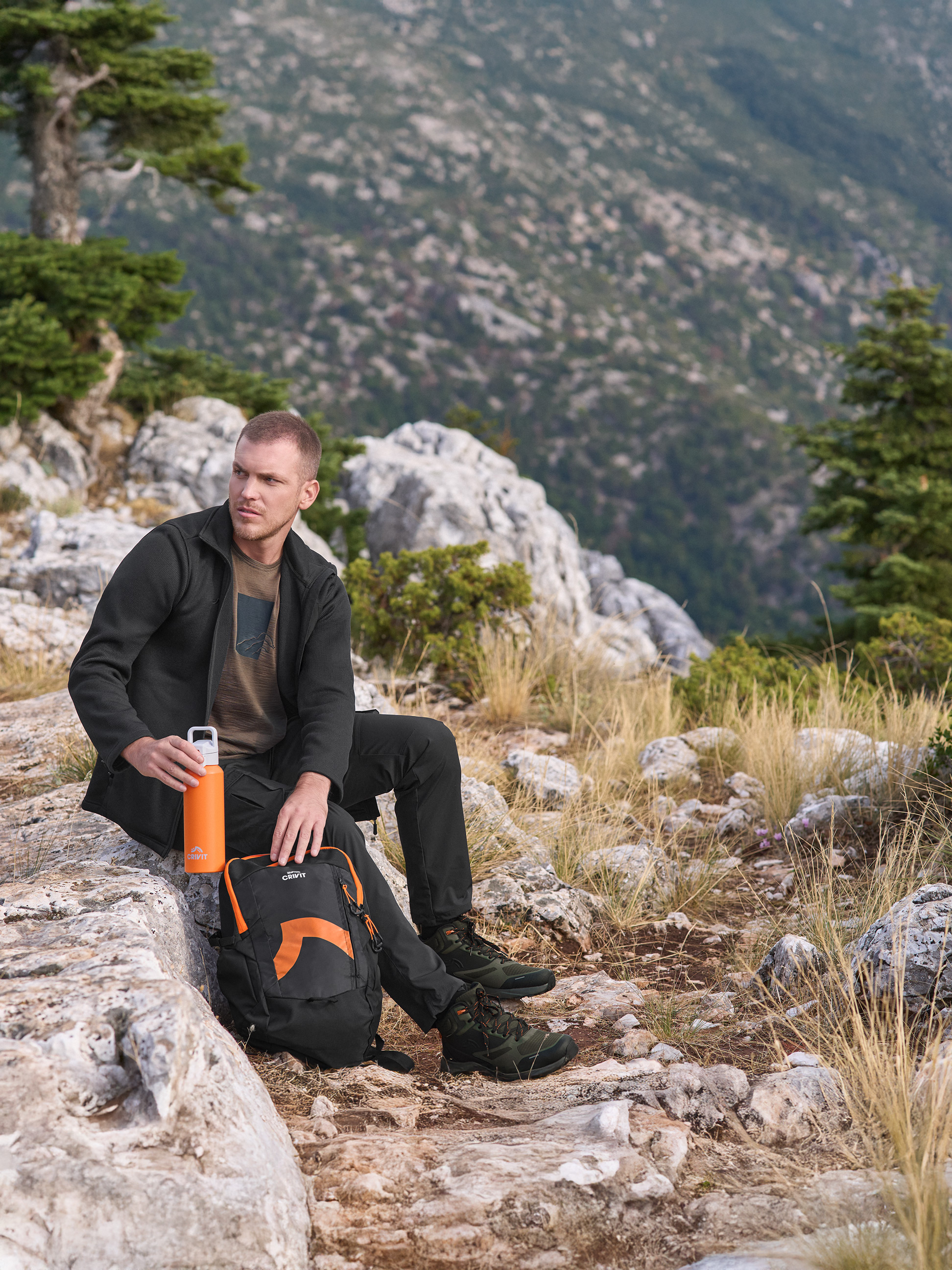 Muž v outdoorovej bunde a nohaviciach Crivit, drží oranžovú fľašu na vodu Crivit, s batohom Crivit.