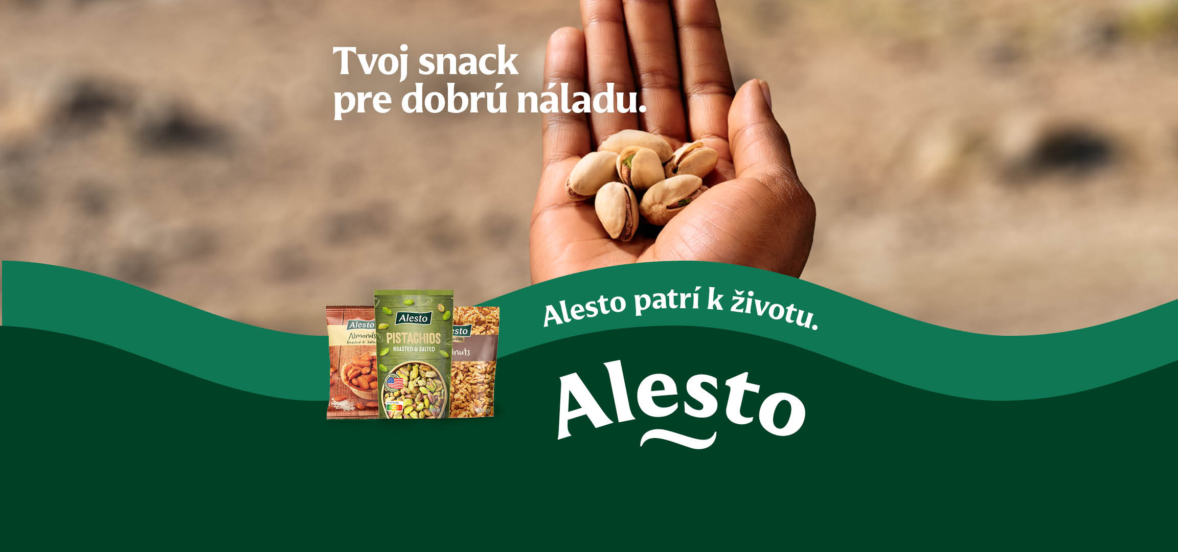 Ruka držiaca pistácie, s baleniami mandlí, pistácií a vlašských orechov. Text: „Tvoj snack pre dobrú náladu. Patrí k životu.“