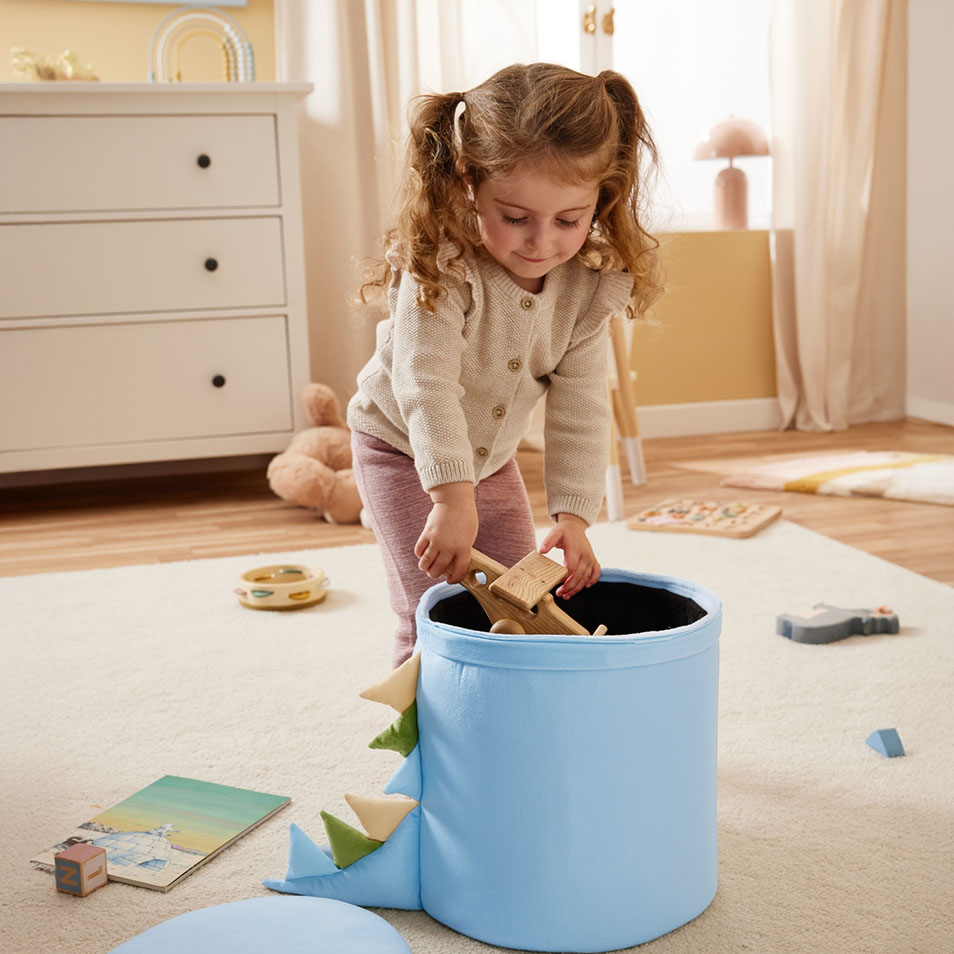 Dieťa vkladá drevené hračky do modrého úložného koša s motívom dinosaura v spálni.