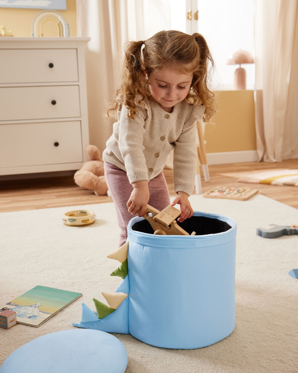 Dieťa vkladá drevené hračky do modrého úložného koša s motívom dinosaura.