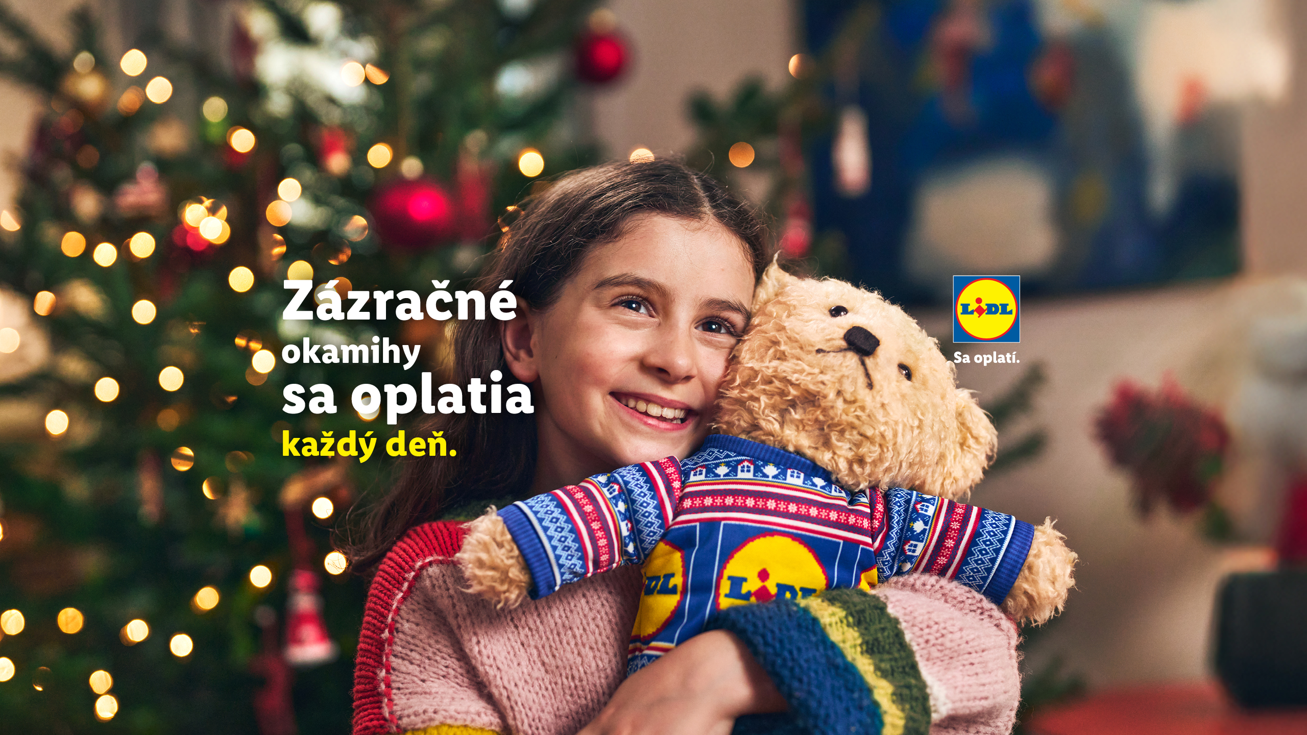 Dievča objíma plyšového medvedíka v Lidl vianočnom svetri pred rozžiareným stromčekom.