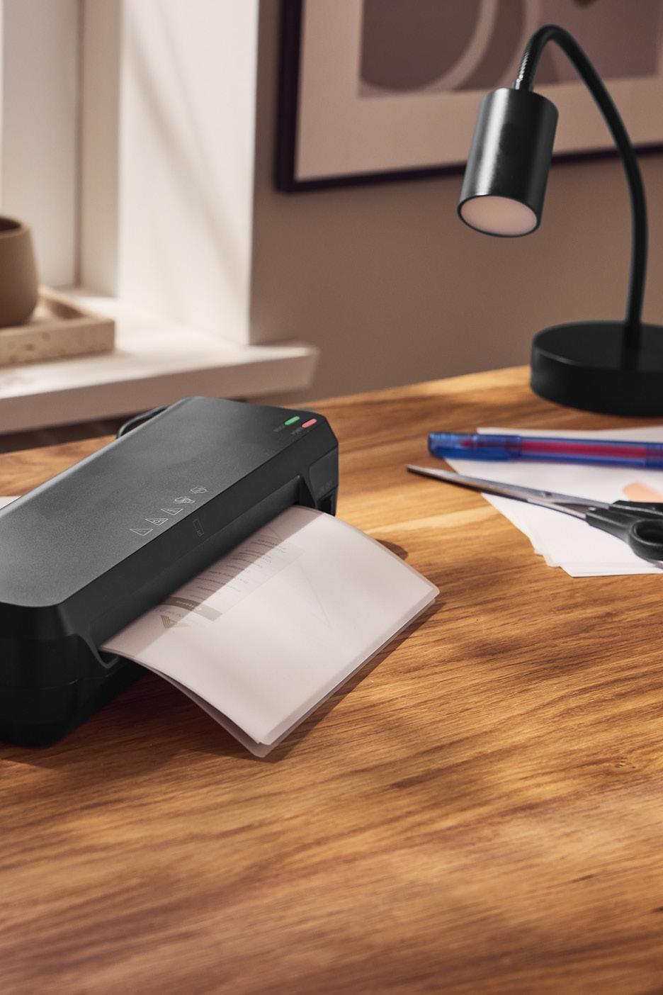 Čierny laminátor laminuje dokument na drevenom stole s lampou a nožnicami.