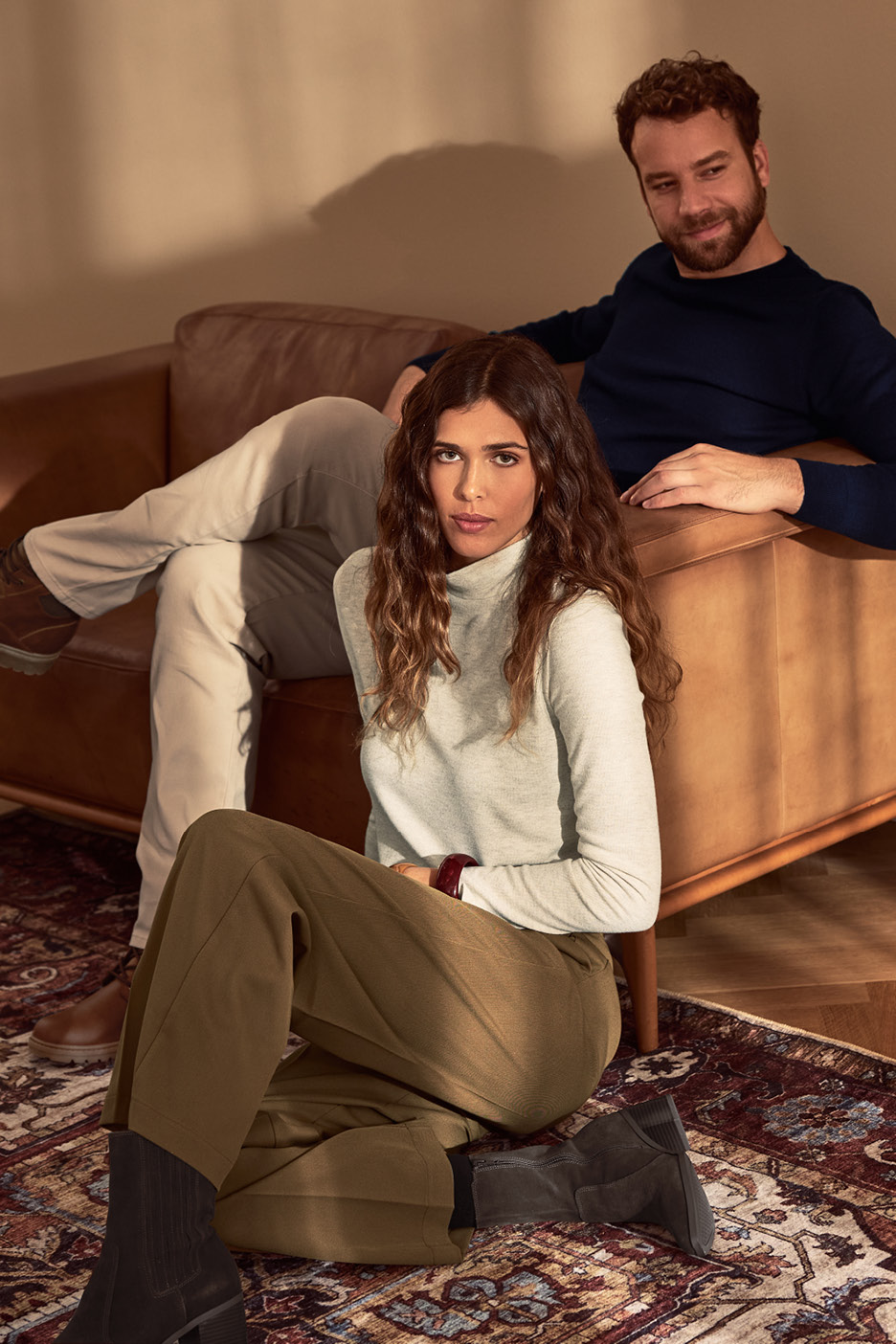 Žena v roláku a nohaviciach sedí na koberci, muž v pozadí na pohovke.