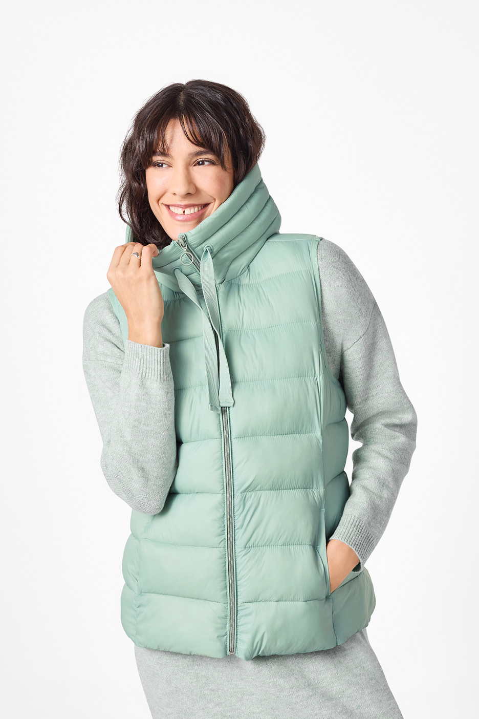 Žena v mätovo zelenej prešívanej veste a sivom svetri.