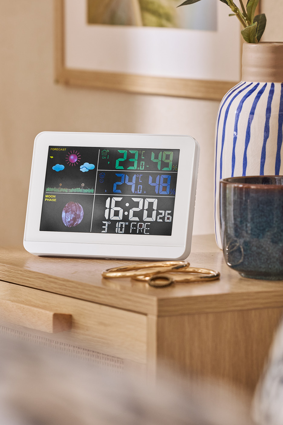 Biely digitálny budík s predpoveďou počasia a fázami mesiaca na drevenom nočnom stolíku.