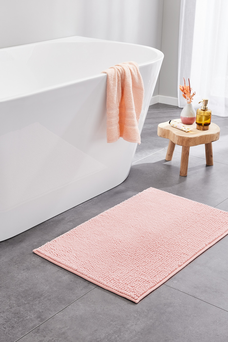 Ružová kúpeľňová predložka a uterák v kúpeľni s bielou vaňou a drevenou stoličkou.