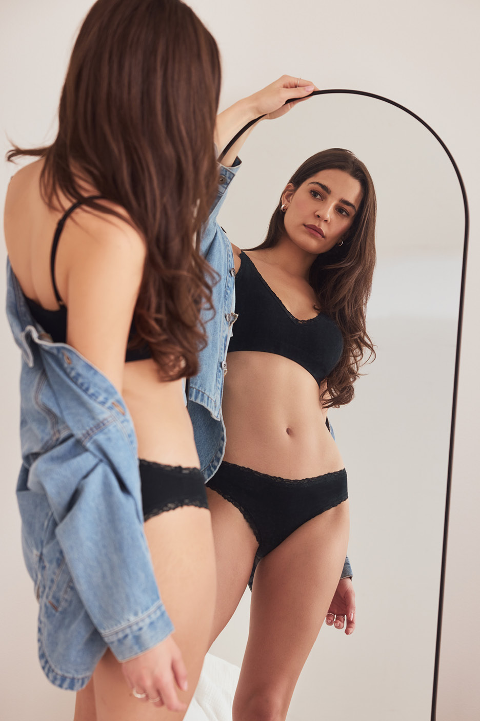 Žena v čiernej čipkovanej podprsenke a nohavičkách sa pozerá do zrkadla, má na sebe džínsovú bundu.