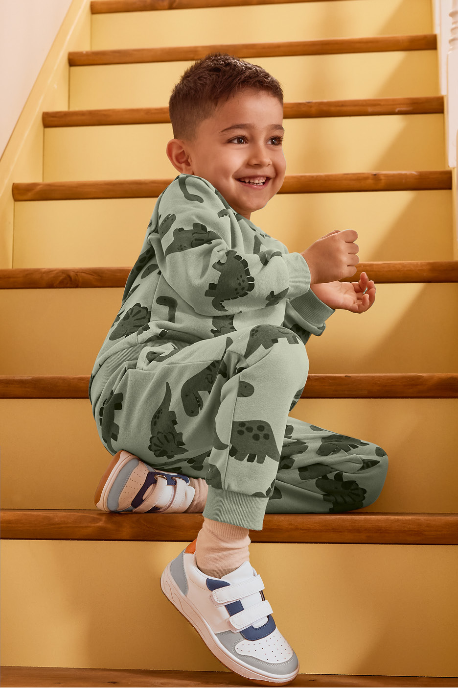 Chlapec v zelenom dinosaurom teplákovom komplete sedí na schodoch a usmieva sa.