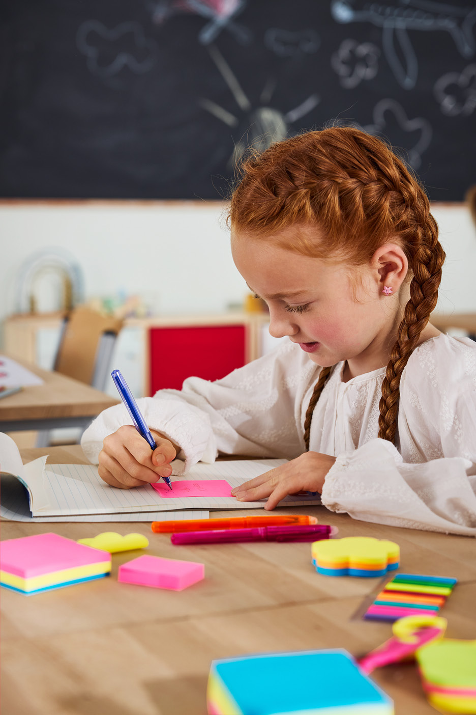 Dievča s ryšavými vlasmi píše perom na ružový poznámkový blok, na stole sú zvýrazňovače.